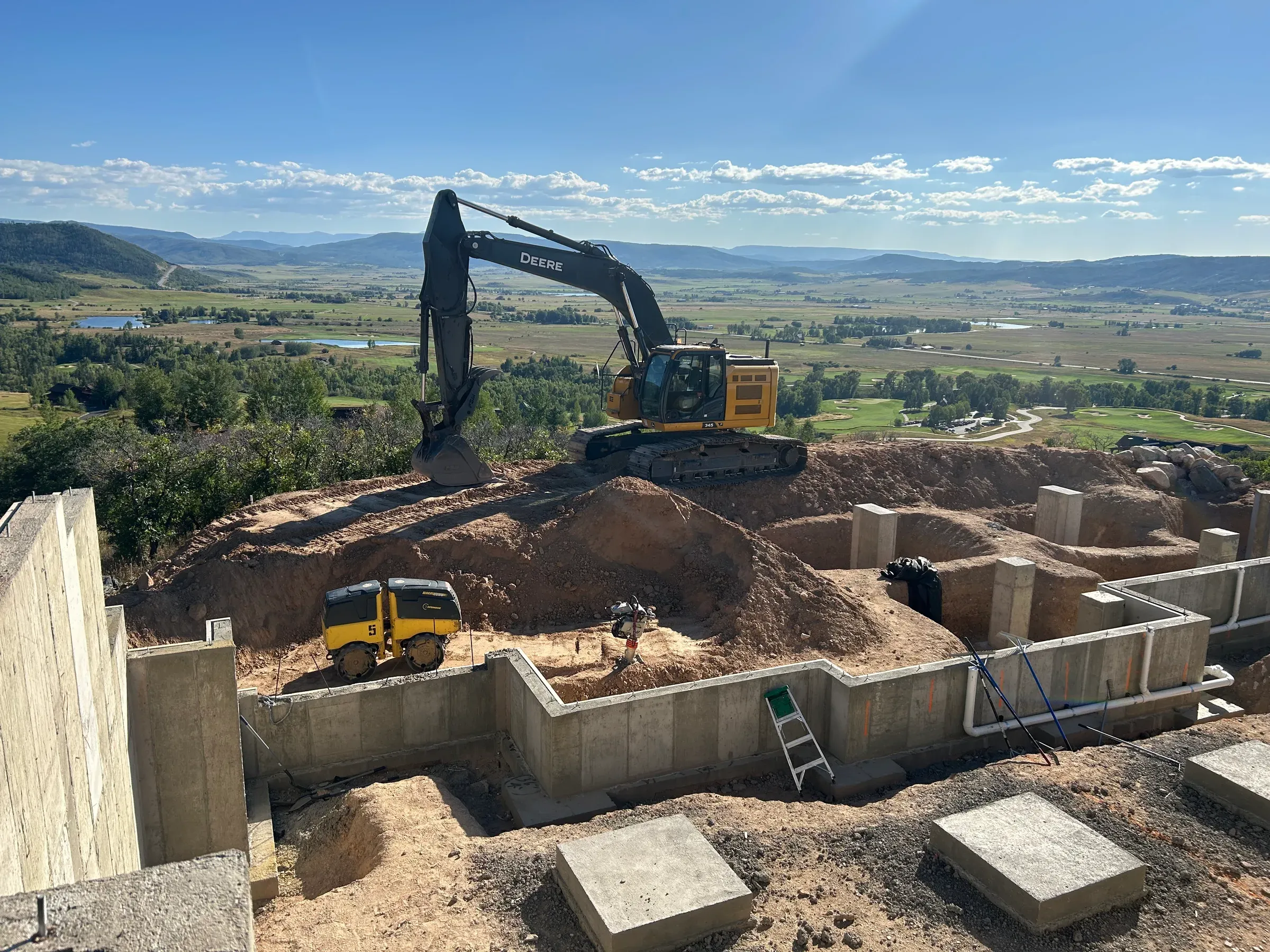 Alpine Earthworks excavator on a hilltop home-foundation site above the high-country valley